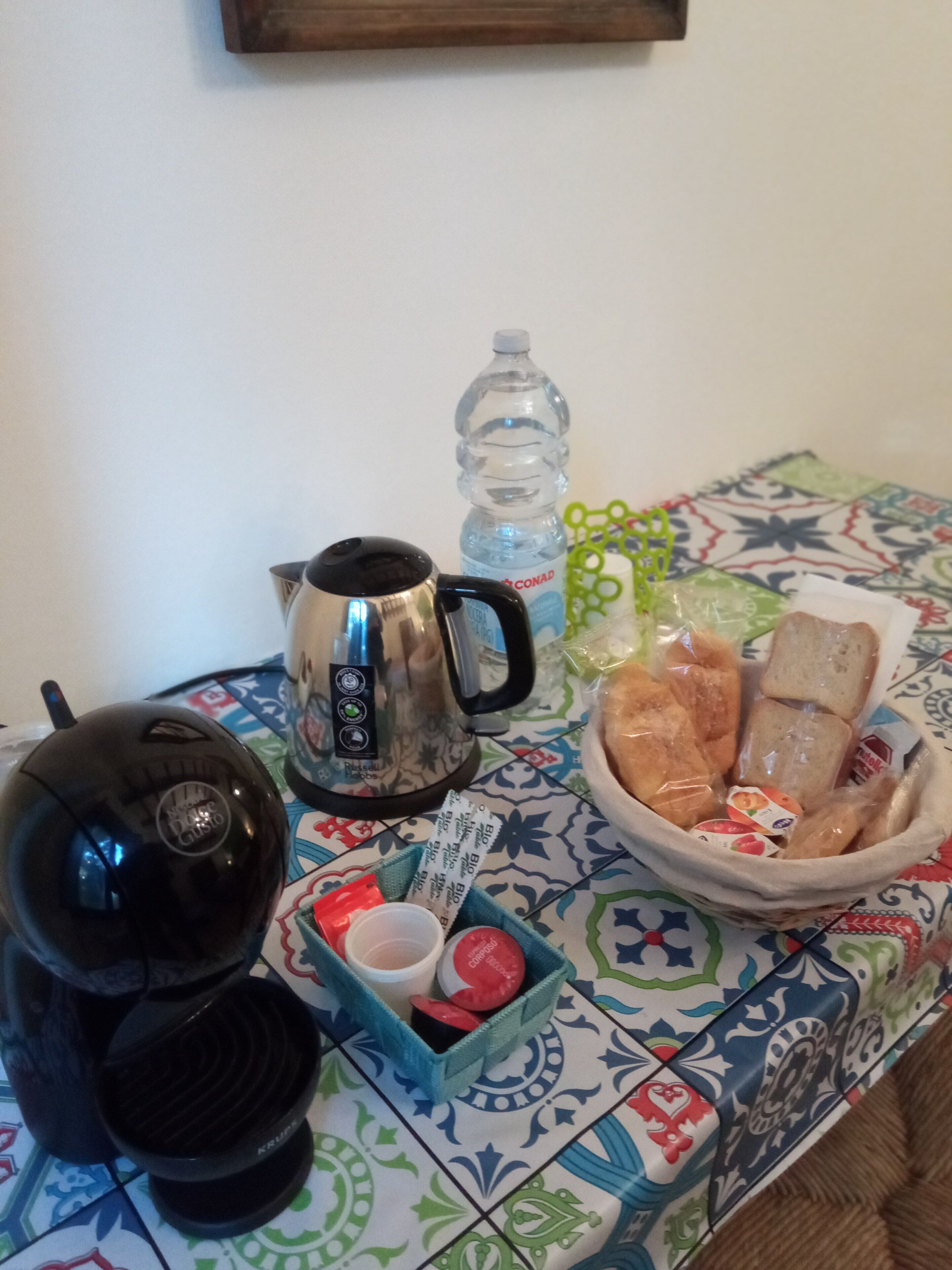 A table with a coffee maker, a kettle, a water bottle, and a basket of bread and snacks.