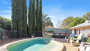 A swimming pool with a curved edge, surrounded by a gravel area and a metal fence. There are lounge chairs and a small table with a tablecloth. Tall trees and a building are visible in the background. - Great Divide Motor Inn (East Toowoomba)