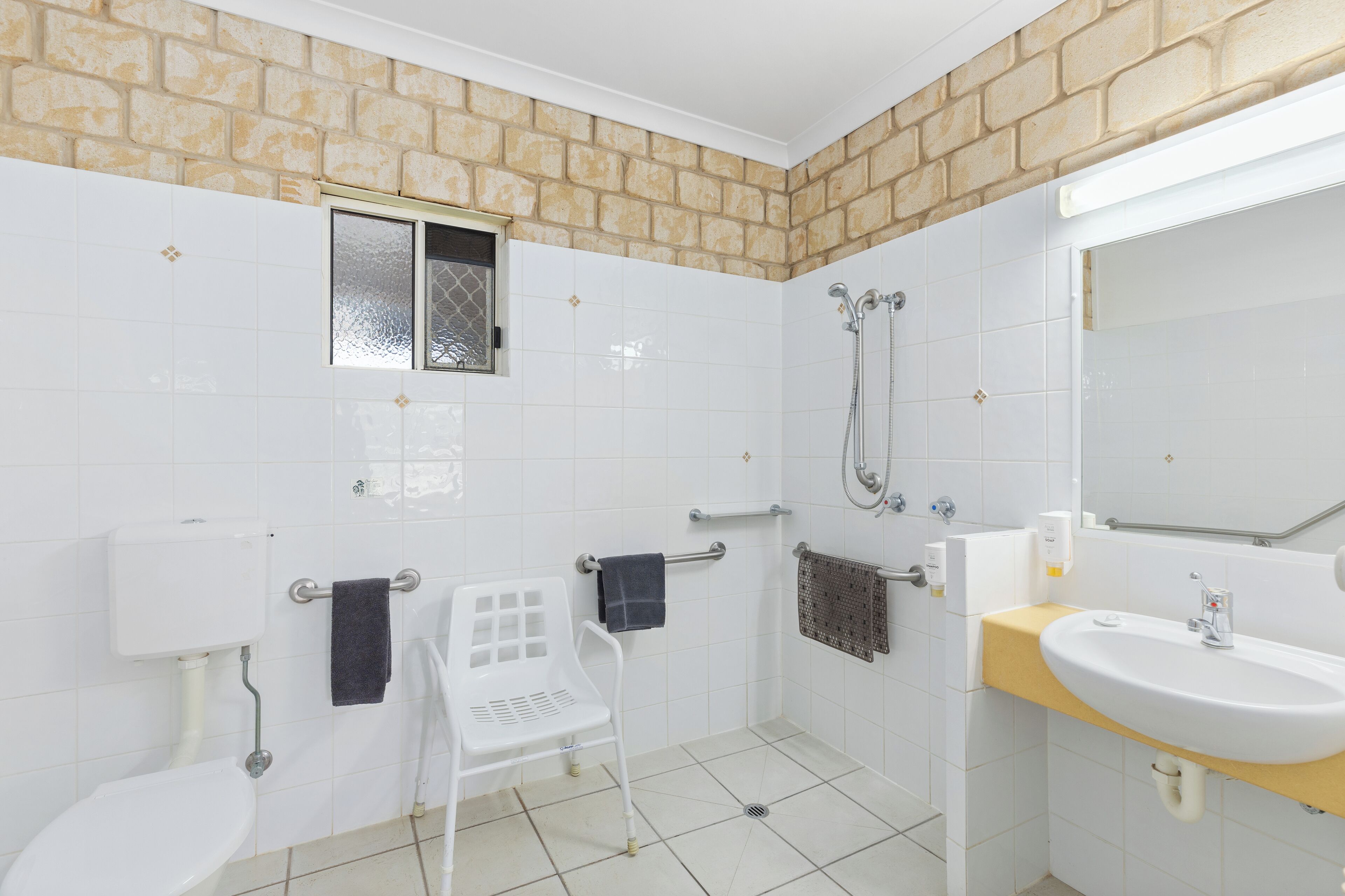 Une salle de bain avec des carreaux blancs, un mur en briques de couleur beige, des toilettes, un lavabo, une douche et une fenêtre.