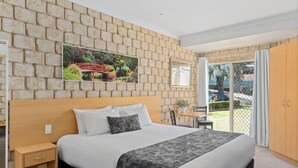 A hotel room with a brick-patterned wall, a large bed with white bedding, a grey patterned throw, a wooden nightstand, a small table with a chair, and a sliding glass door leading to an outdoor area. - Great Divide Motor Inn (East Toowoomba)