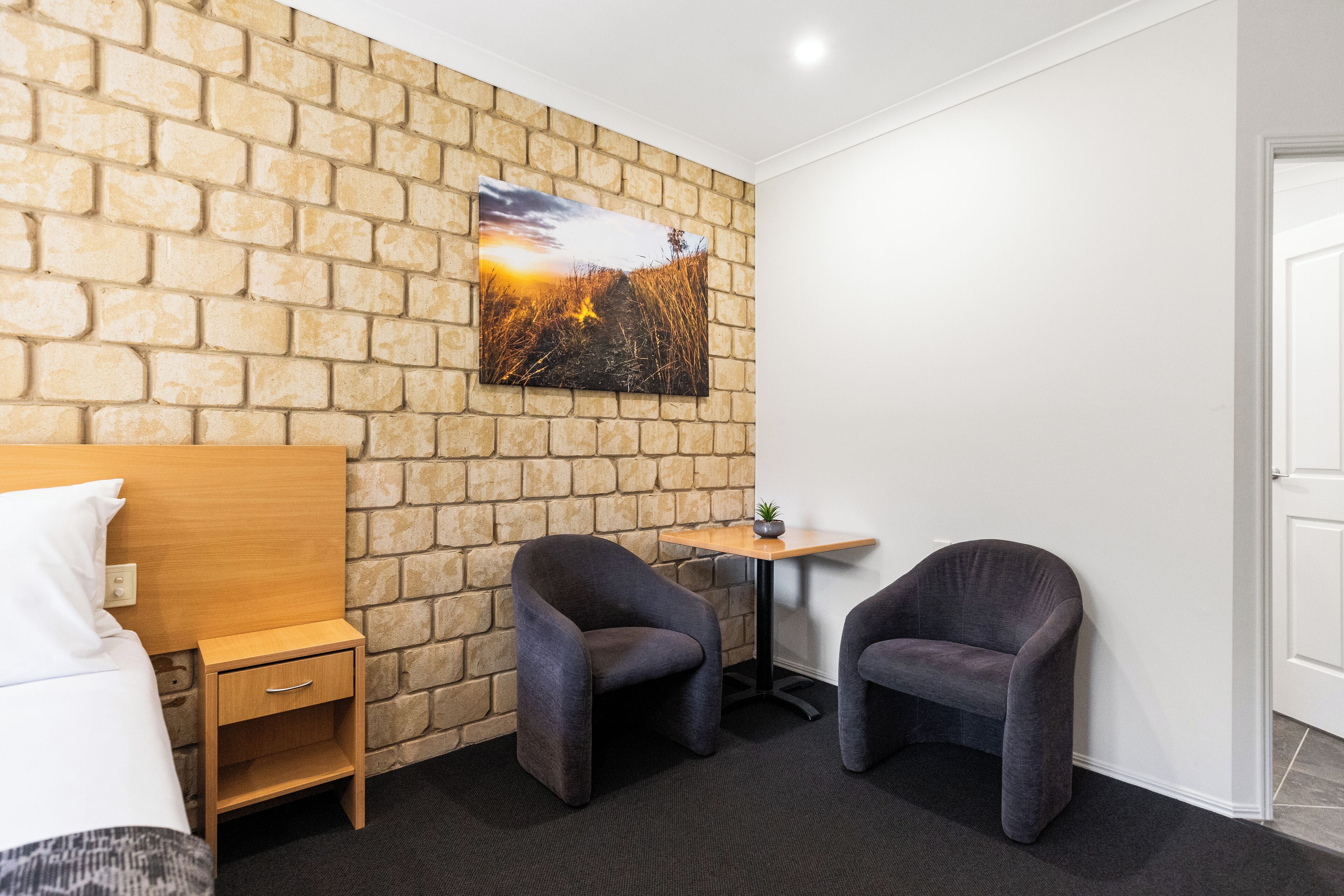 Habitación de hotel con pared de ladrillo, cama con ropa blanca, dos sillones oscuros, una mesita de madera con una planta y un cuadro de un atardecer.