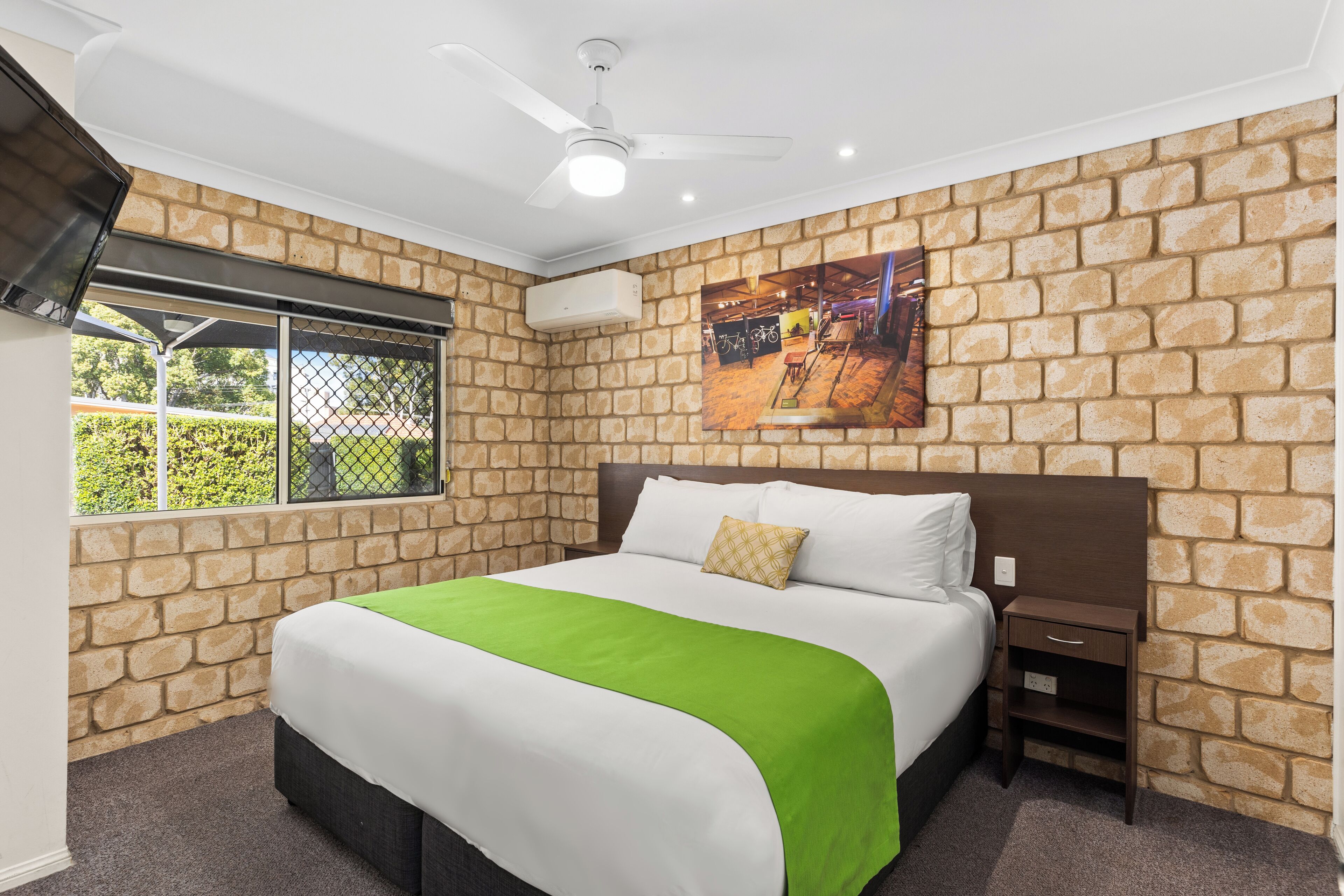 Una camera da letto con una parete a mattoni, un letto con copripiumino verde e bianco, un comodino e una ventola a soffitto.