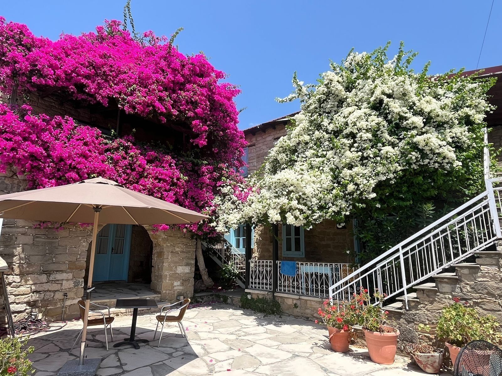 Un patio de piedra con una mesa y sillas, una escalera y un edificio cubierto de enredaderas rosadas y blancas.