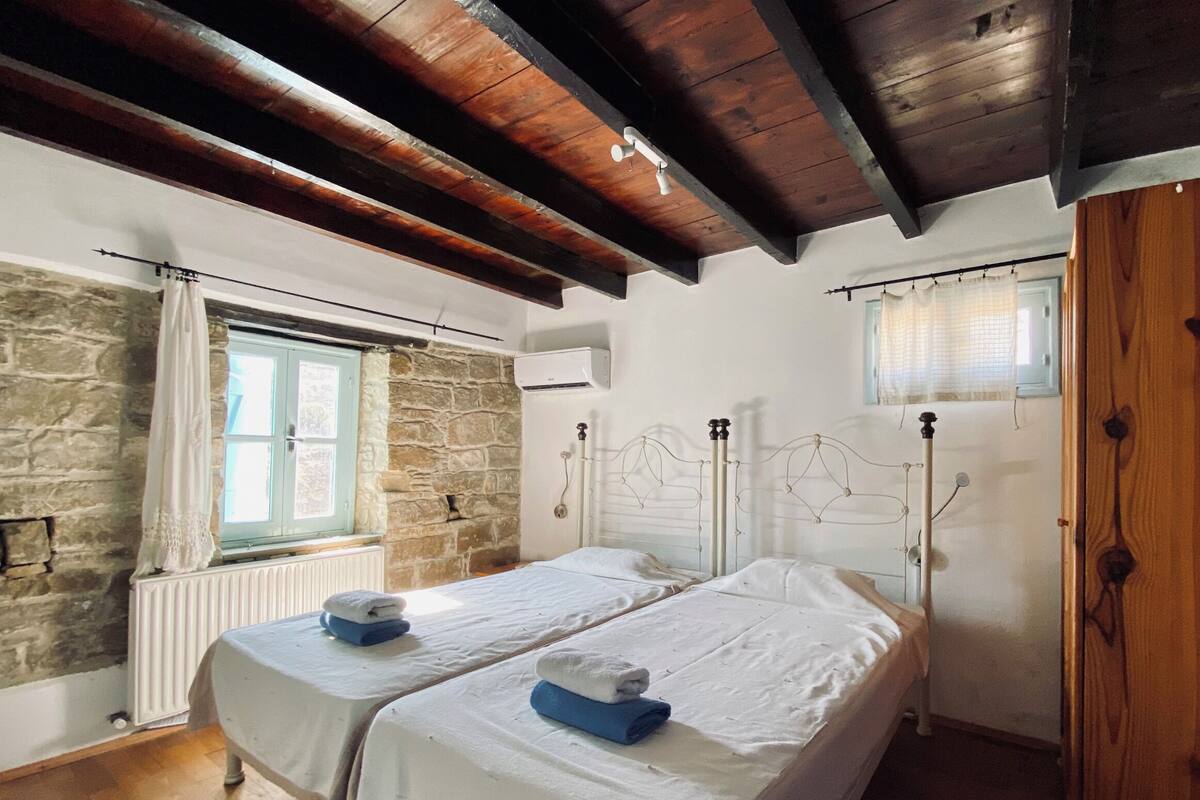 A room with two beds, stone walls, wooden beams, a window with curtains, and an air conditioning unit.