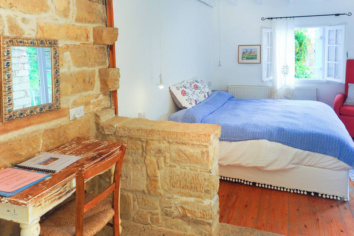 A bedroom with a stone wall, a wooden desk with papers, a bed with a blue and white bedspread, and a red chair.