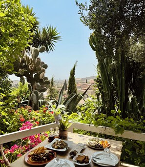 Mesa al aire libre con diversos platos, rodeada de abundante vegetación y cactus, con vistas a un paisaje urbano.