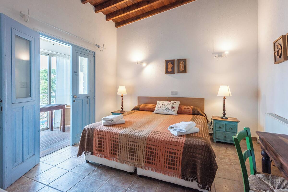 A bedroom with a bed, two bedside tables with lamps, a chair, a wooden bench, and a door leading to an outdoor area.