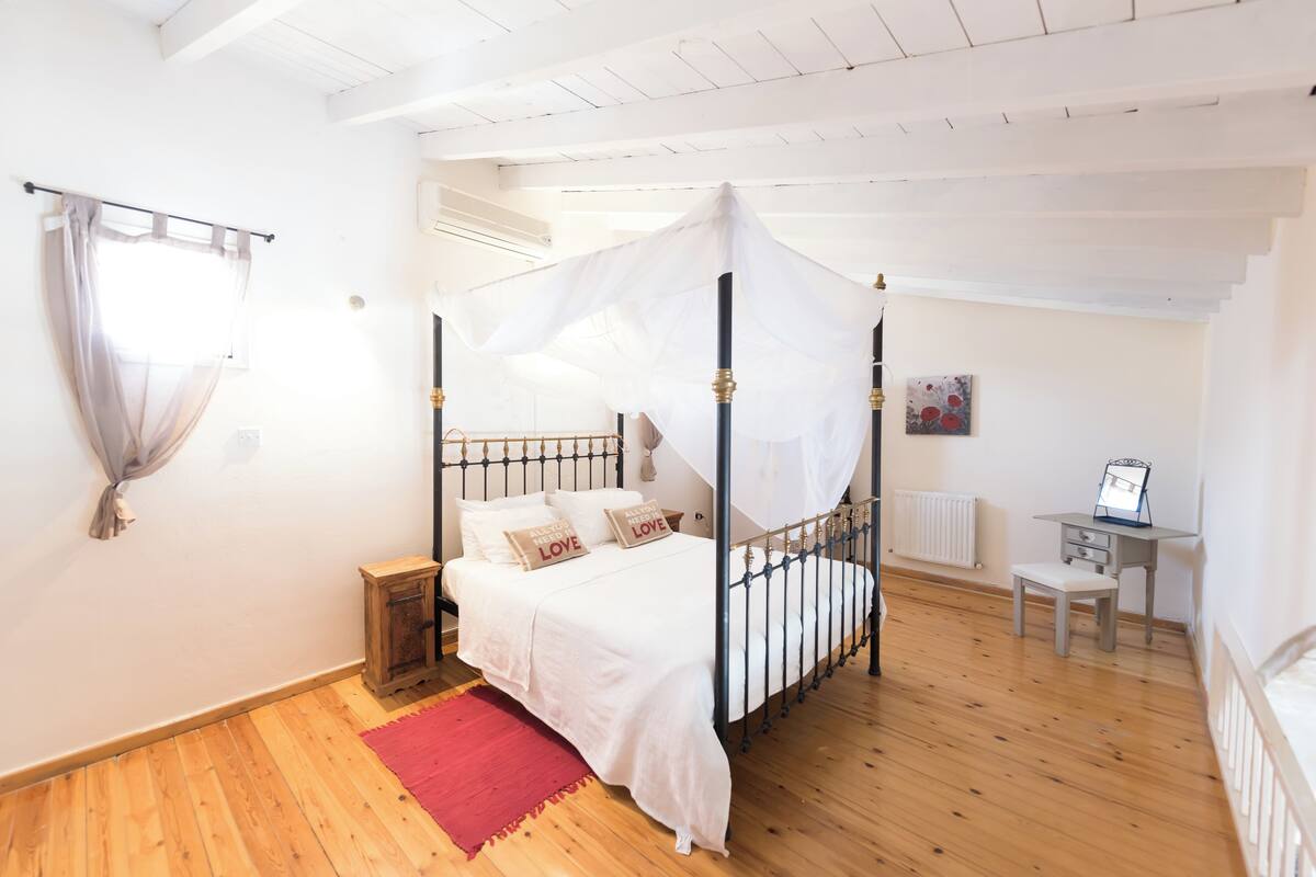 A bedroom with a four-poster bed, a small bedside table, a red rug, and a desk with a chair.