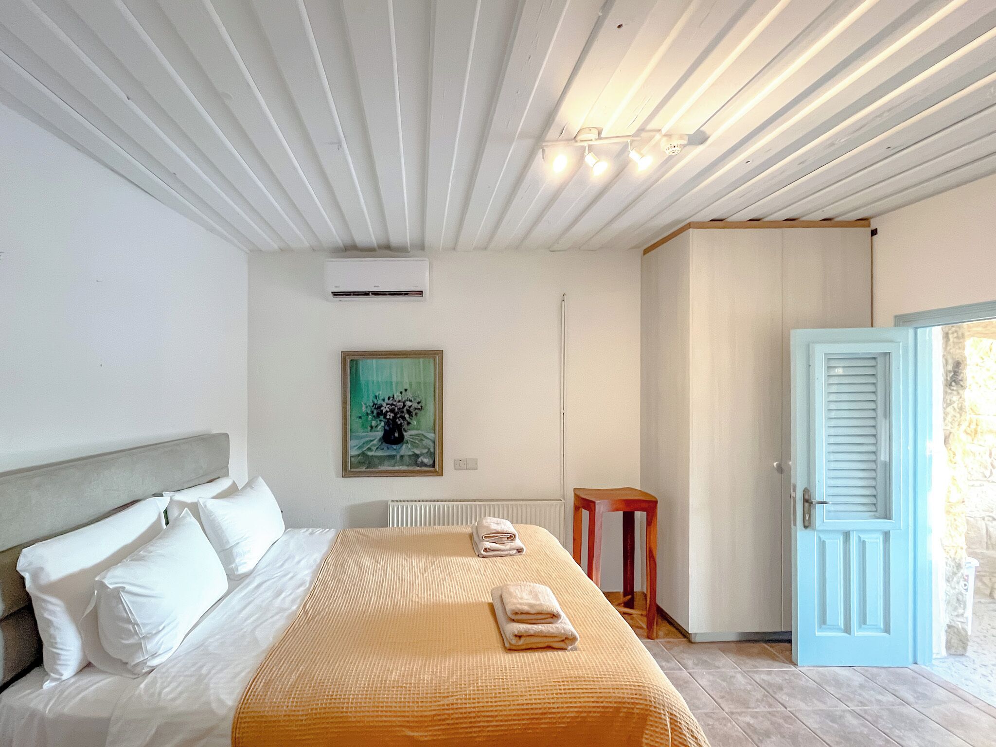 A hotel room with a bed, a painting on the wall, an air conditioning unit, and a door with a stone wall outside.