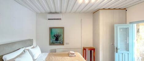 A hotel room with a bed, a painting on the wall, an air conditioning unit, and a door with a stone wall outside.
