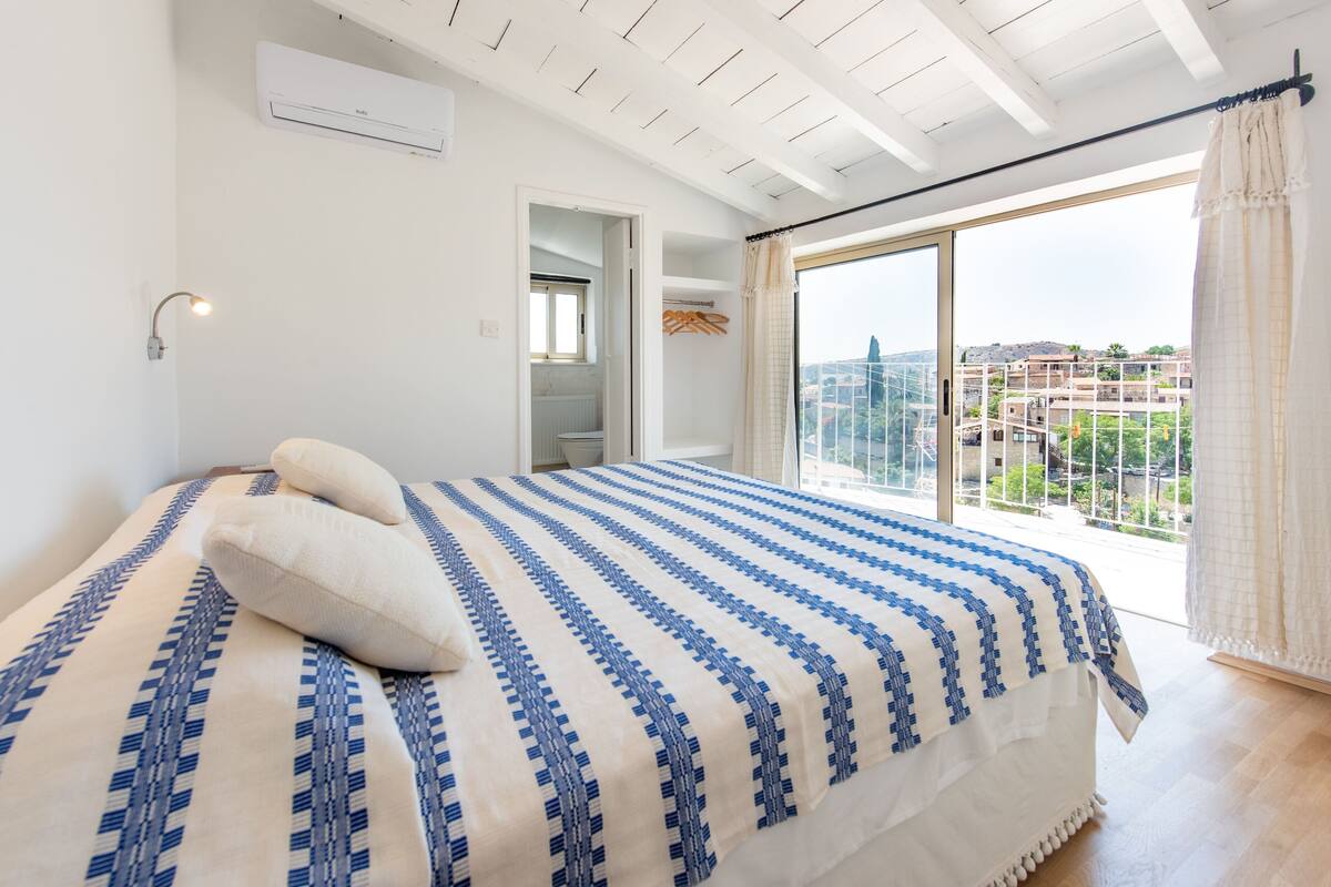 A bedroom with a large bed, a window with a view of buildings, and a ceiling fan.