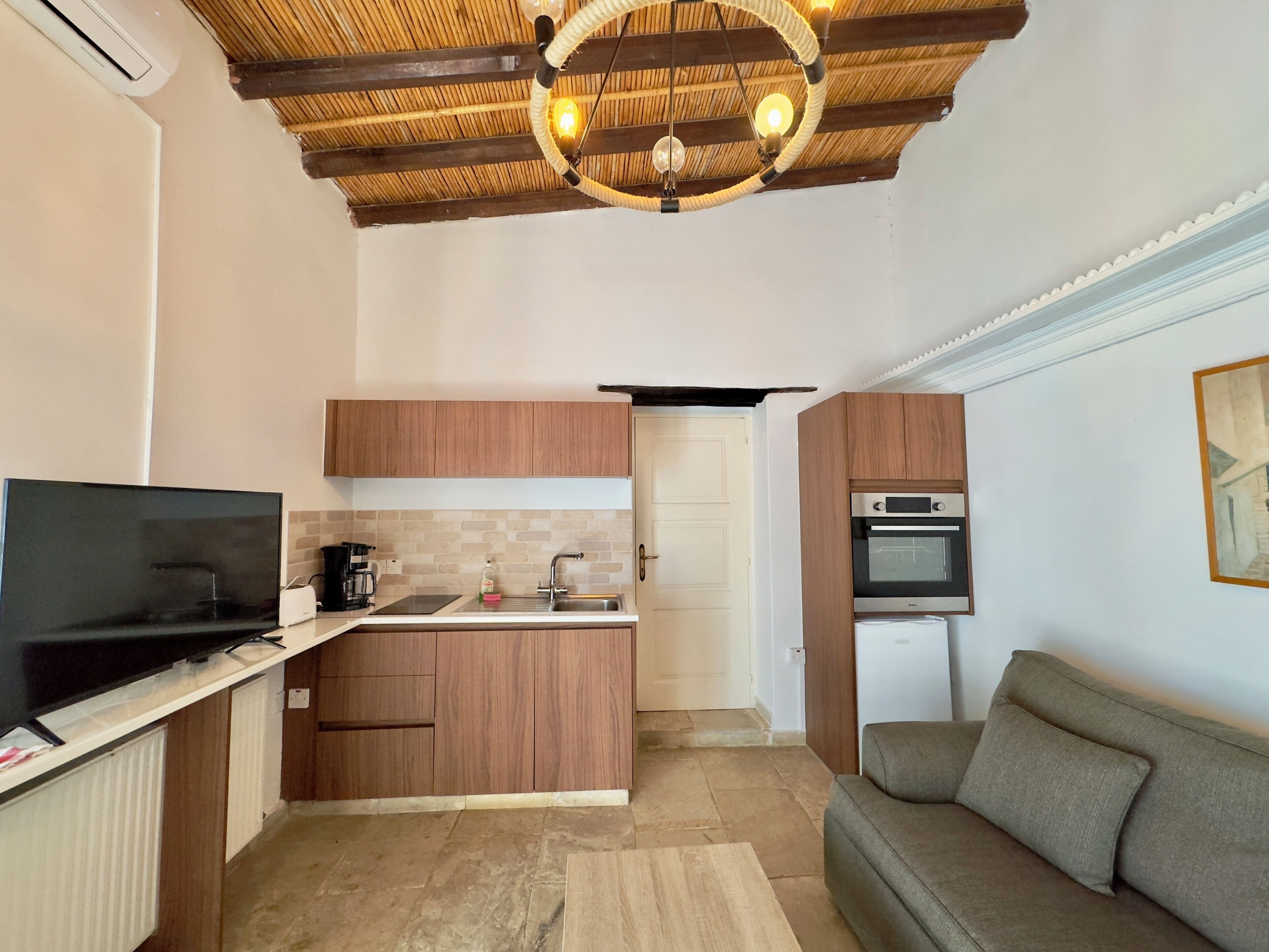 A compact living space with a kitchenette, a television, a sofa, and a ceiling with exposed wooden beams and a hanging light fixture.