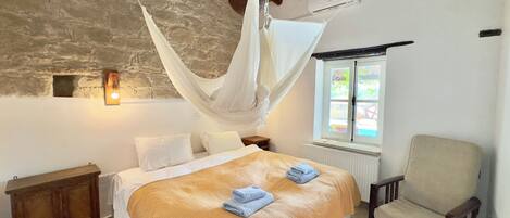 A bedroom with a bed, a chair, a nightstand, a window, and a stone wall.