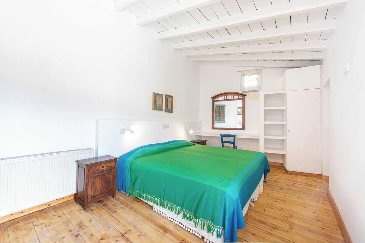 A bedroom with a wooden floor, a bed with a green bedspread, a wooden nightstand, a chair, a wardrobe, and a mirror.