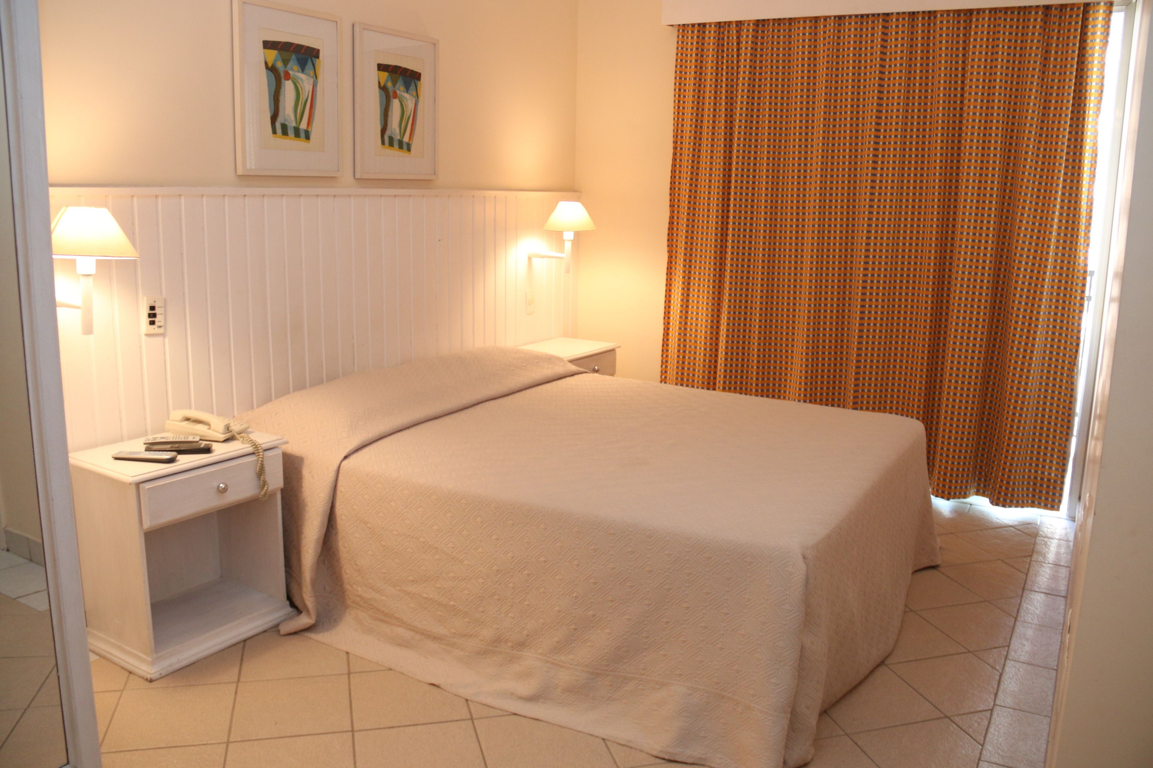 Un lit bien fait dans une chambre d’hôtel, avec une tête de lit blanche, deux tables de chevet avec des lampes et une fenêtre avec des rideaux.