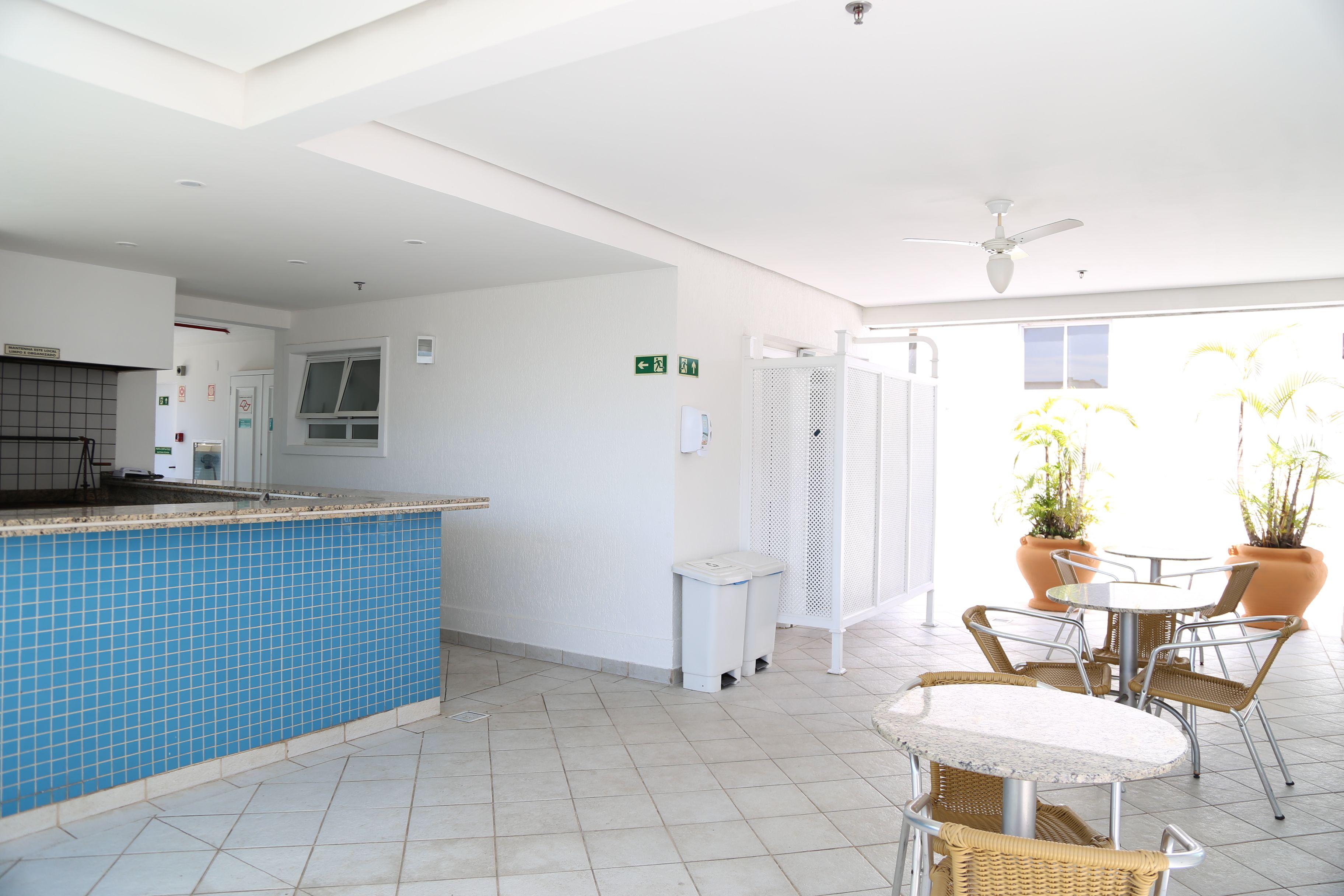Una cocina limpia y moderna con salpicadero de azulejos azules, encimera blanca y ventilador de techo. Hay una pequeña zona de estar con sillas de mimbre y una mesa, además de plantas en macetas al fondo.