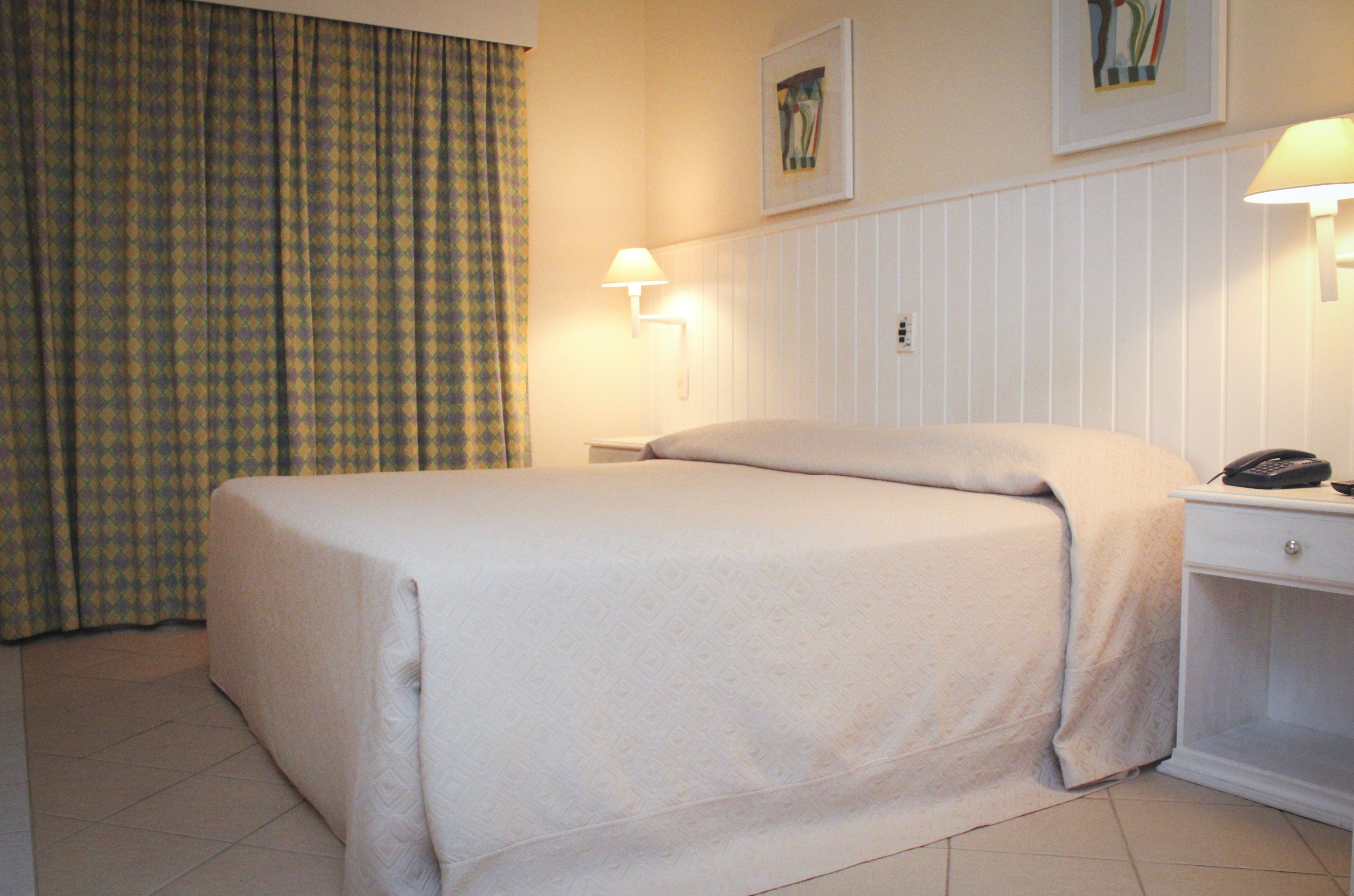 Habitación de hotel con cama, mesita de noche, lámpara, teléfono y cortina.