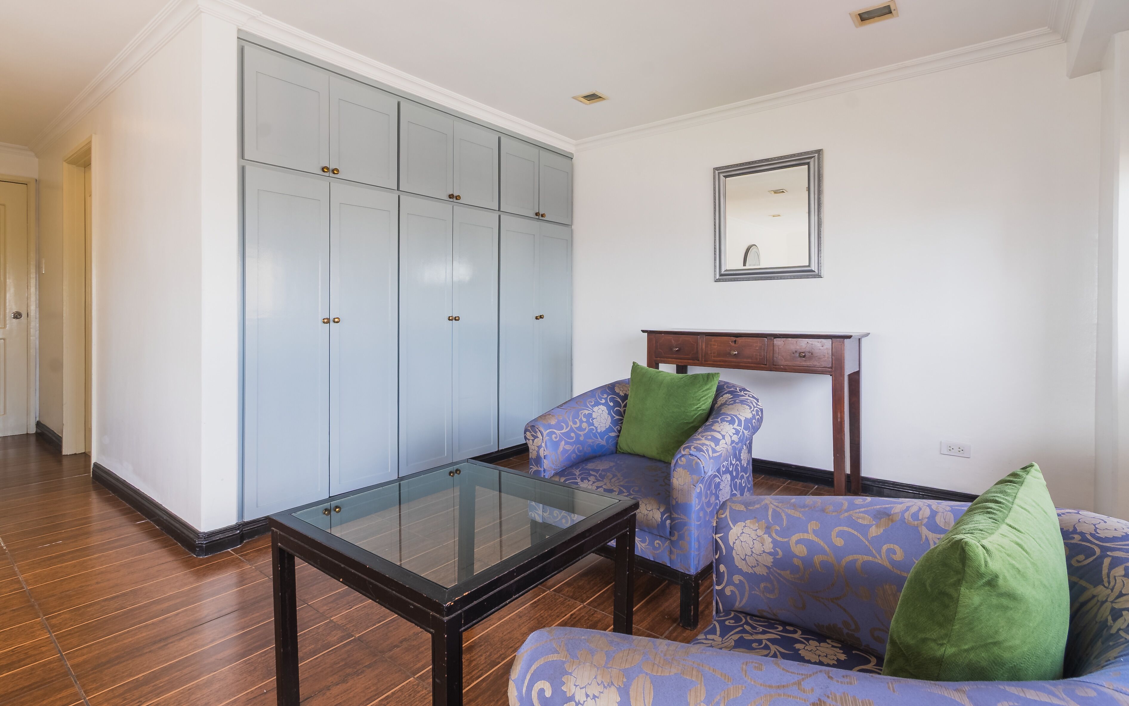 A room with a glass-top table, a patterned armchair, a blue wardrobe, and a wooden nightstand.