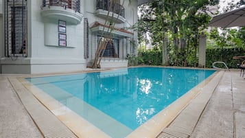 A swimming pool with a tiled edge, surrounded by a concrete deck, and a white building with balconies and metal railings.