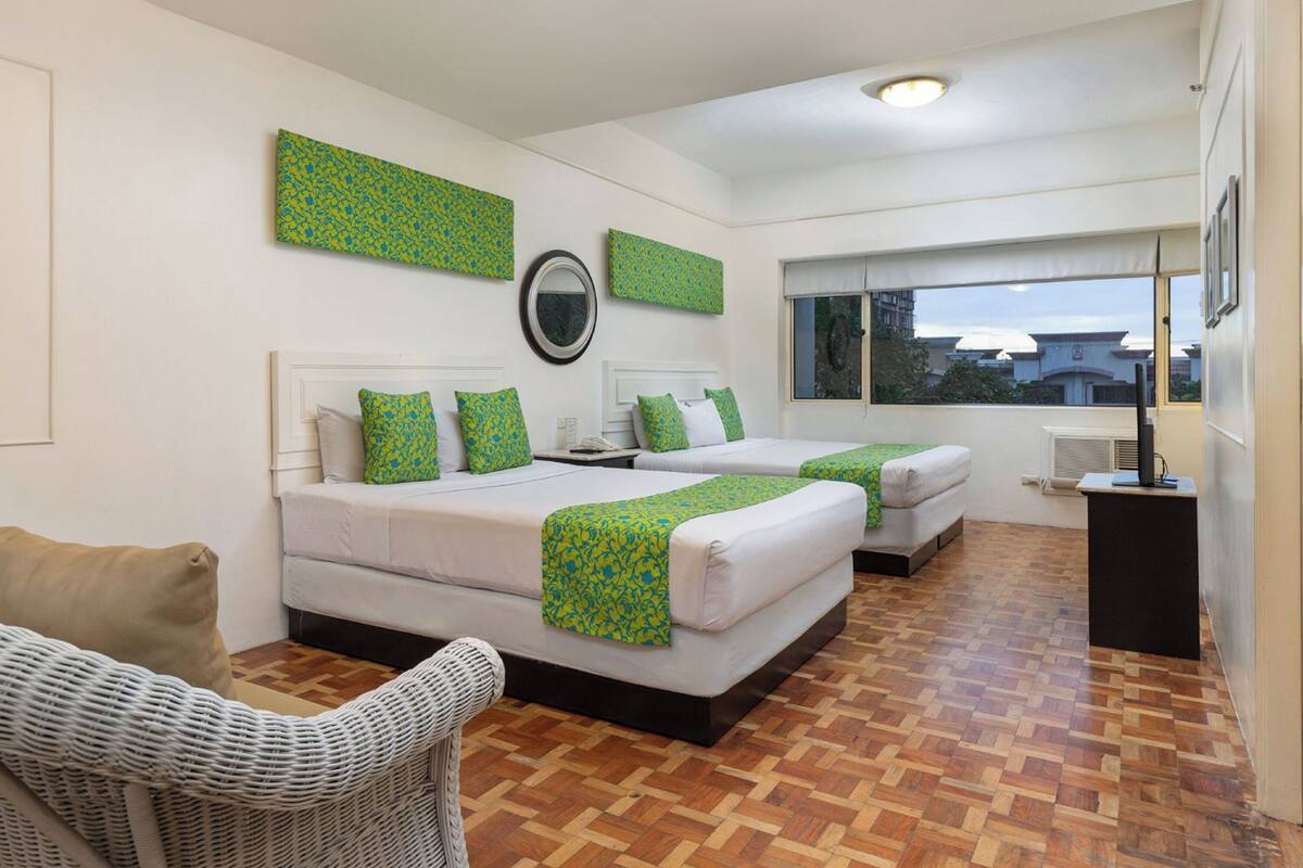 A hotel room with two beds, a wicker chair, a small table, and a window with a view of buildings.