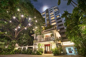 A hotel with a well-lit entrance, surrounded by lush greenery and a large tree with hanging lights. - Orchid Garden Suites (Pasay)