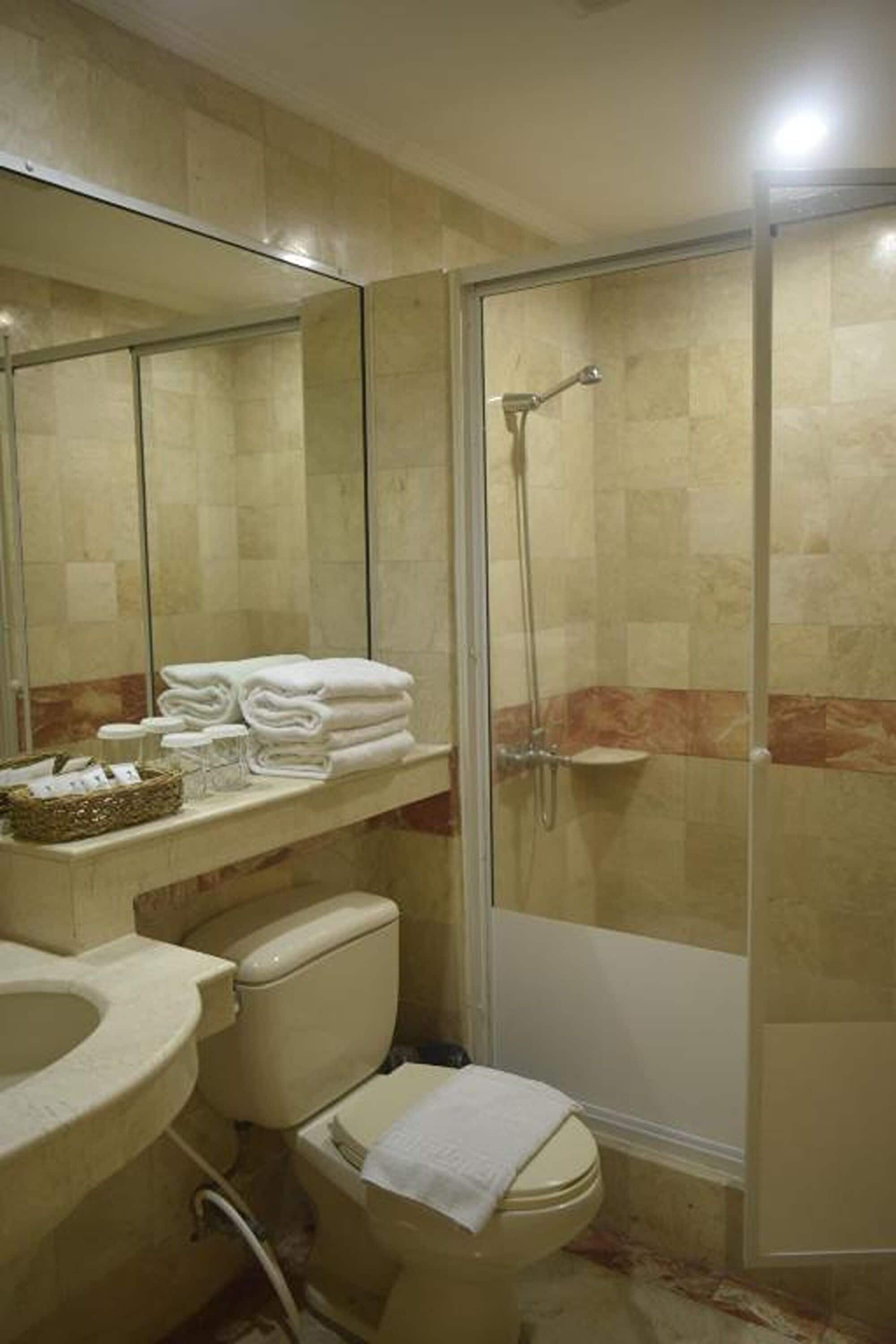A bathroom with a toilet, sink, and shower. There are towels stacked on the sink counter.