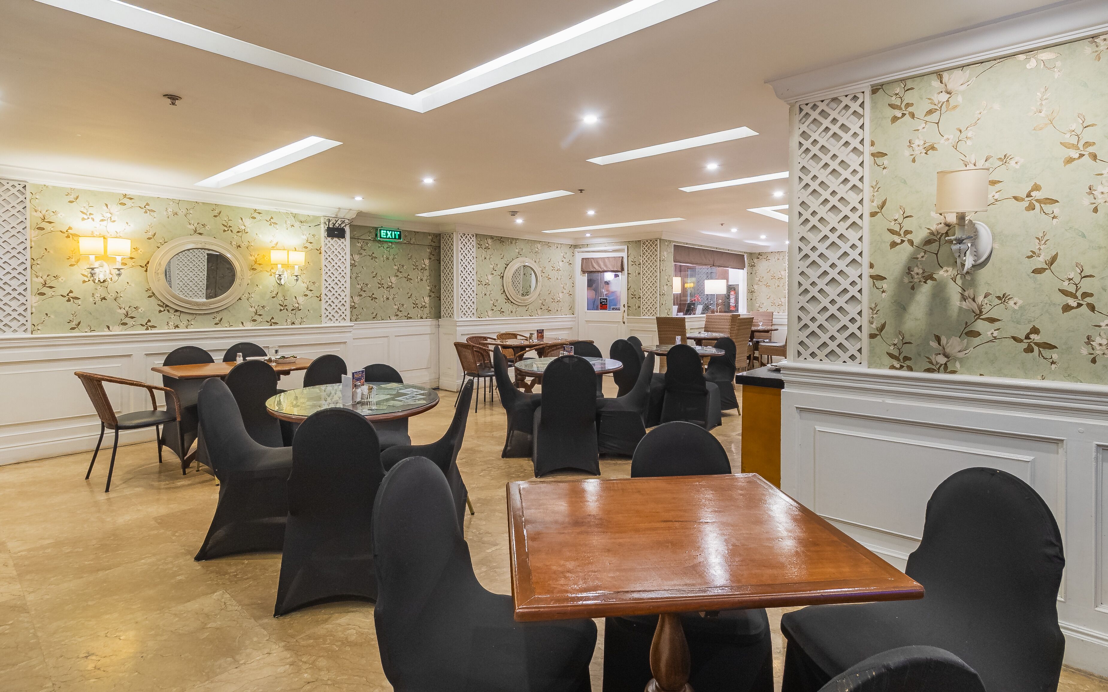 Une salle à manger d’hôtel avec plusieurs tables et chaises, un panneau de sortie bien visible et un papier peint à motifs floraux.