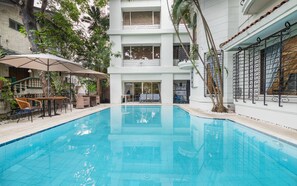 A swimming pool with a clear blue water surface, surrounded by a white building with balconies and a covered outdoor seating area with umbrellas.