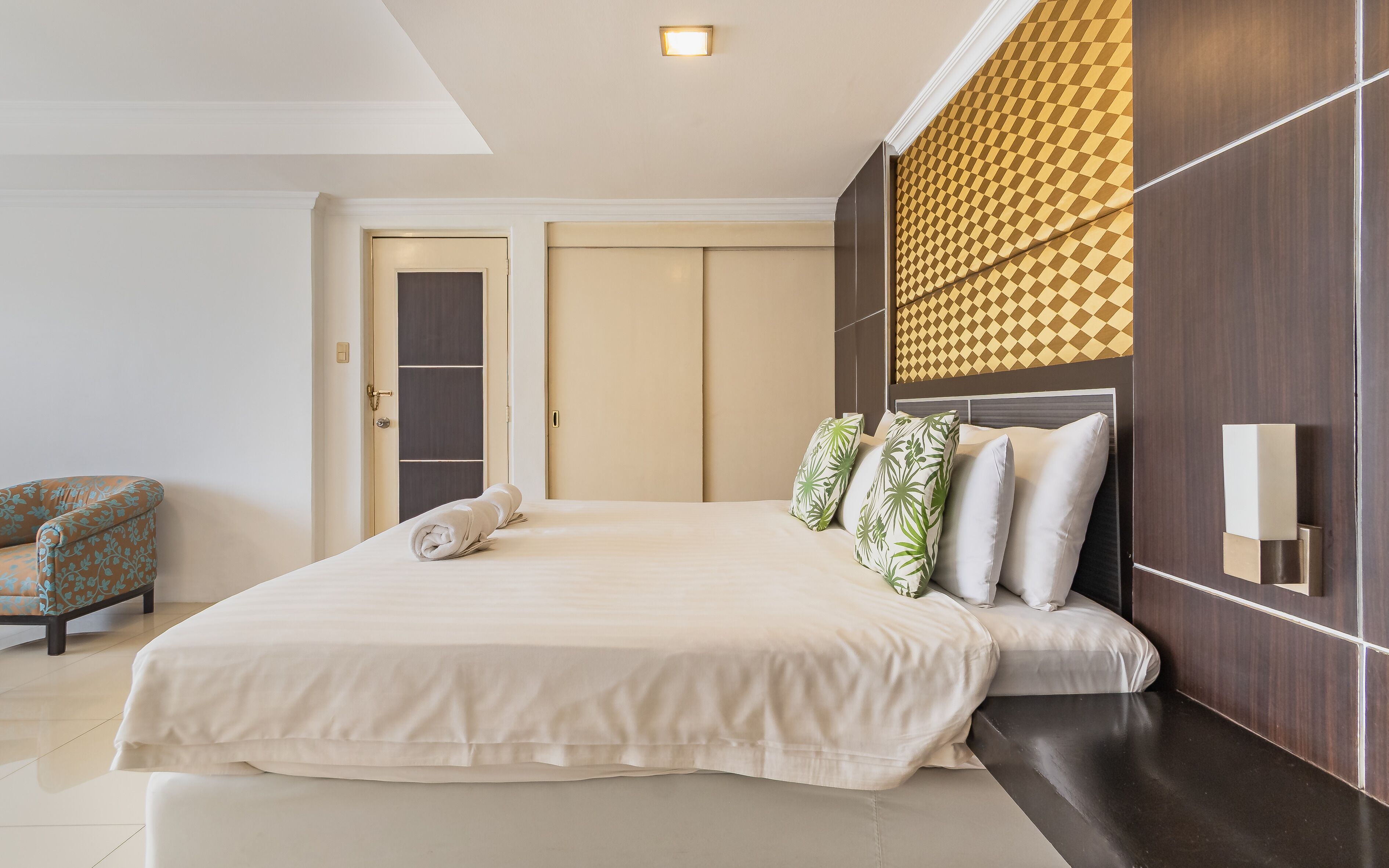 A modern hotel room with a large bed, a seating area with a chair, and a wardrobe.
