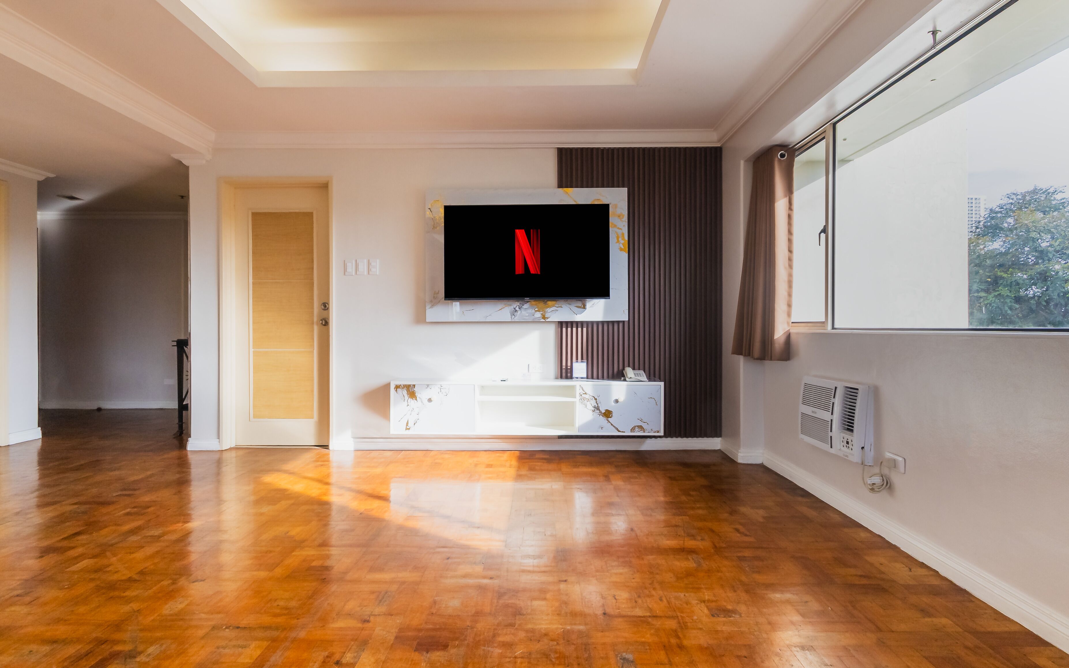 Ein modernes Wohnzimmer mit Flachbildfernseher an der Wand, Holzboden, einer integrierten Unterhaltungseinrichtung und einem großen Fenster mit Vorhängen.