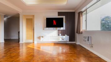 Ein modernes Wohnzimmer mit Flachbildfernseher an der Wand, Holzboden, einer integrierten Unterhaltungseinrichtung und einem großen Fenster mit Vorhängen.