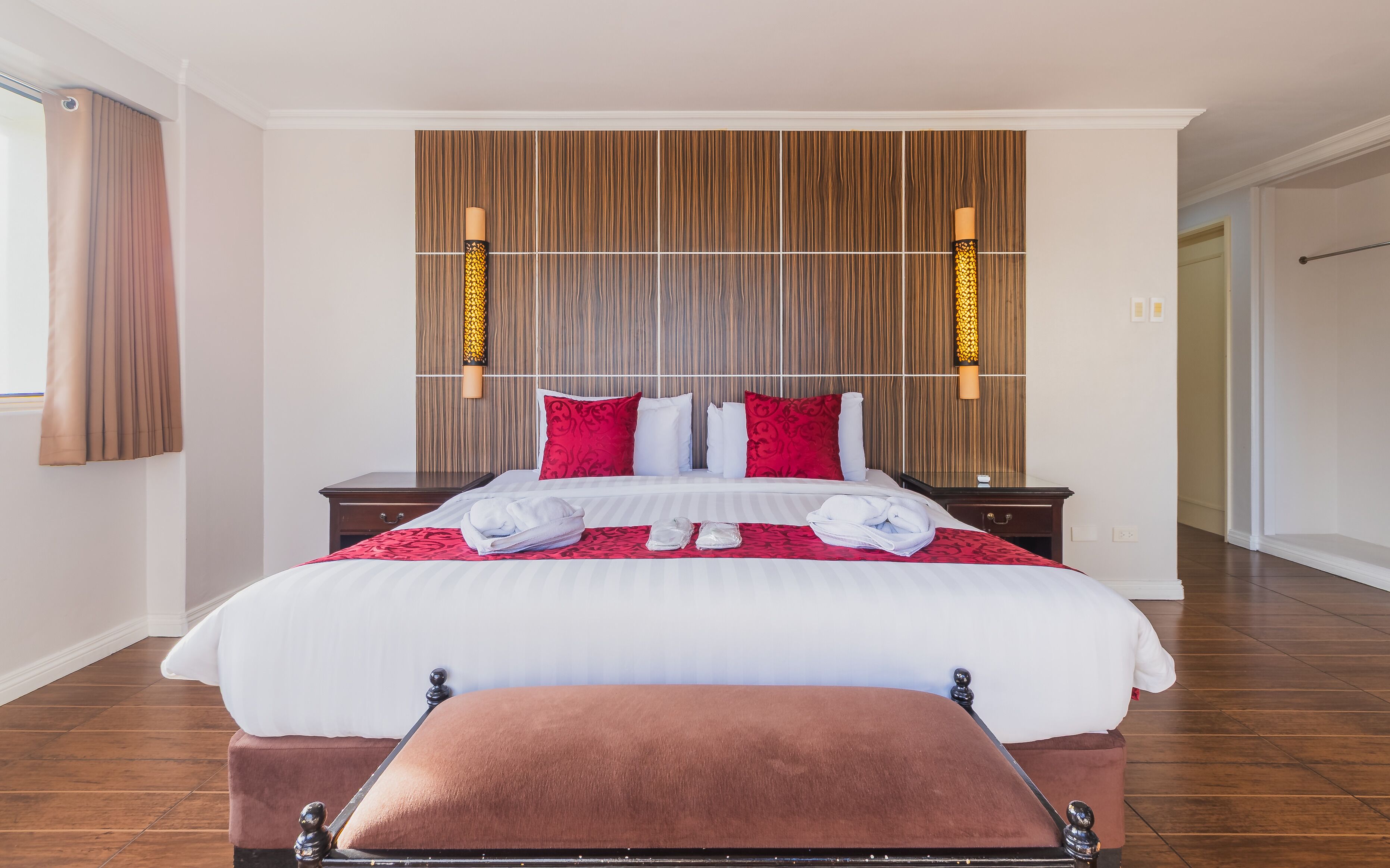 Habitación de hotel con una cama grande, cabecera de madera, un banco marrón y una mesita de noche.