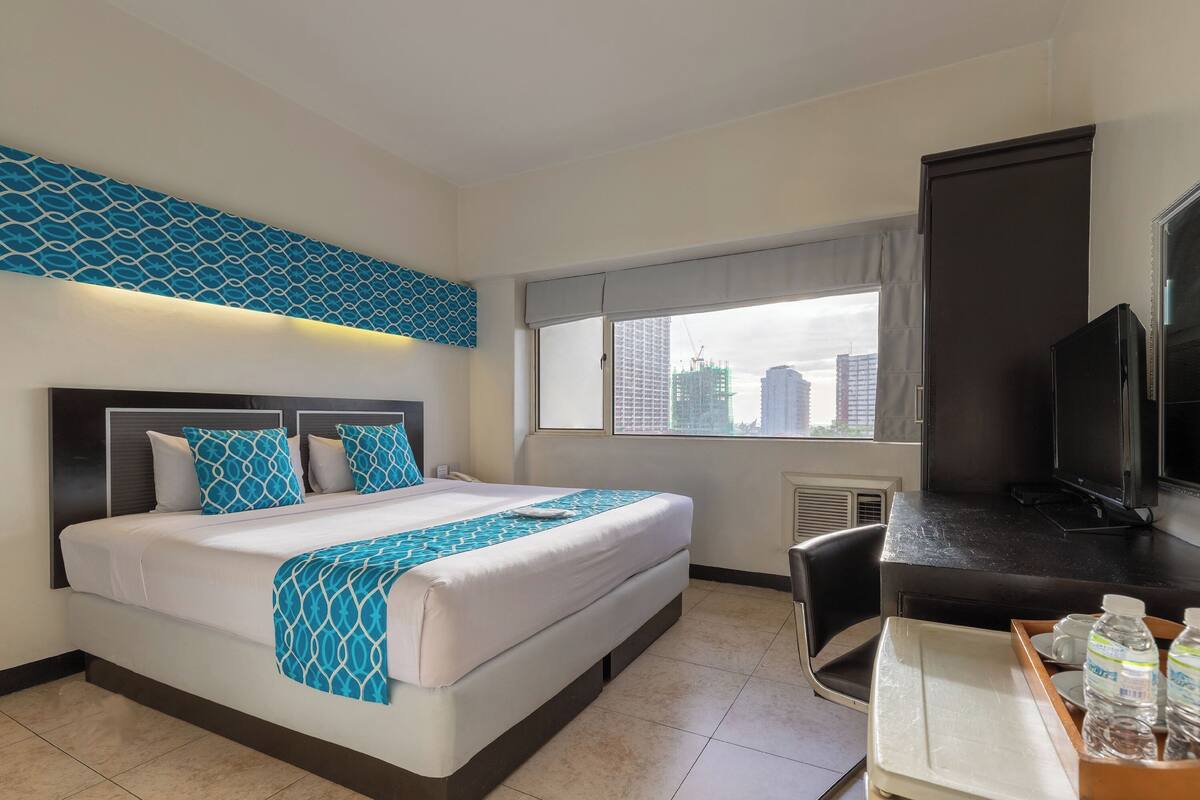 A hotel room with a large bed, a desk with a TV, and a window with a city view.