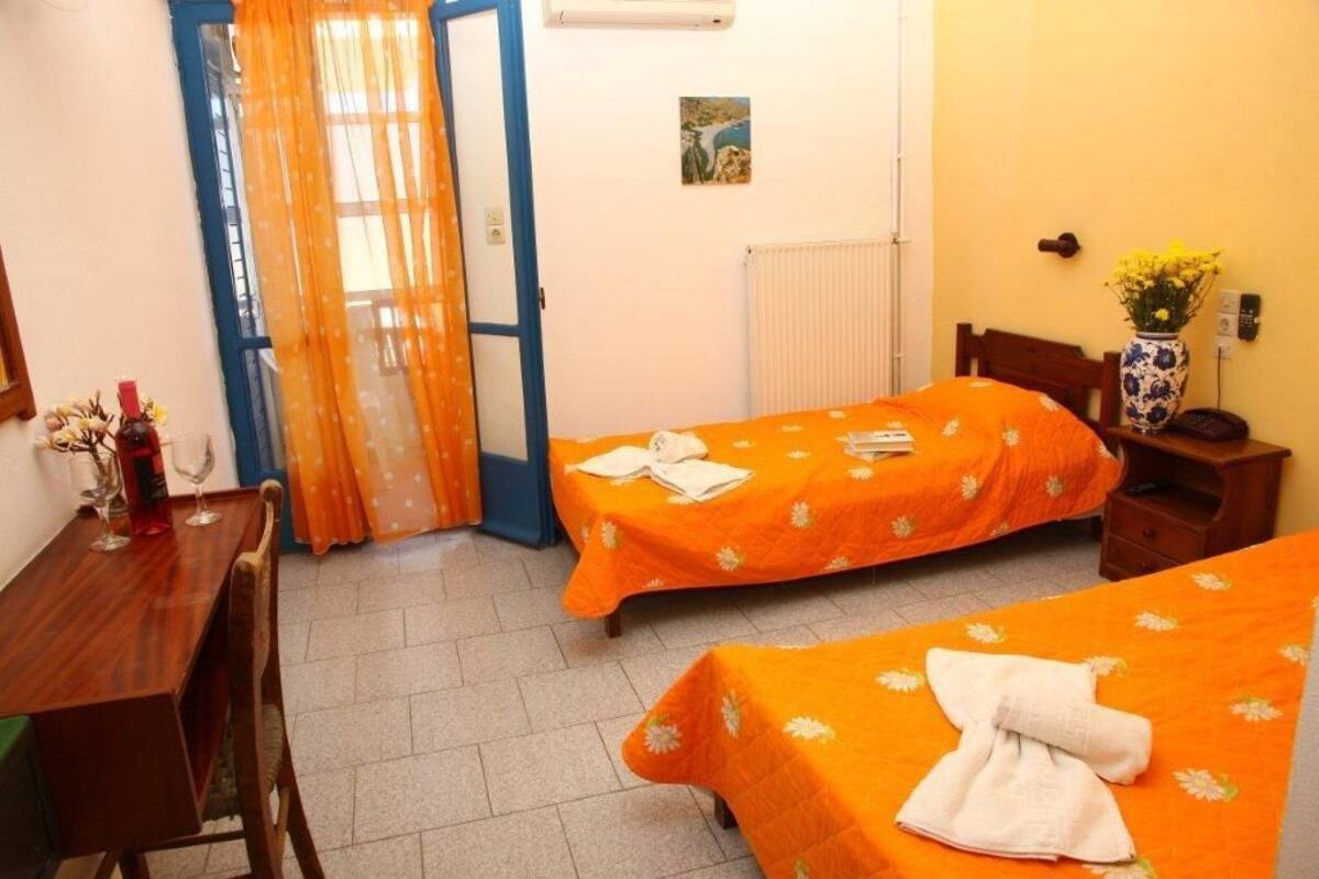 A room with two beds, a wooden desk, a chair, a radiator, a small table with a vase of flowers, and a door with orange curtains.