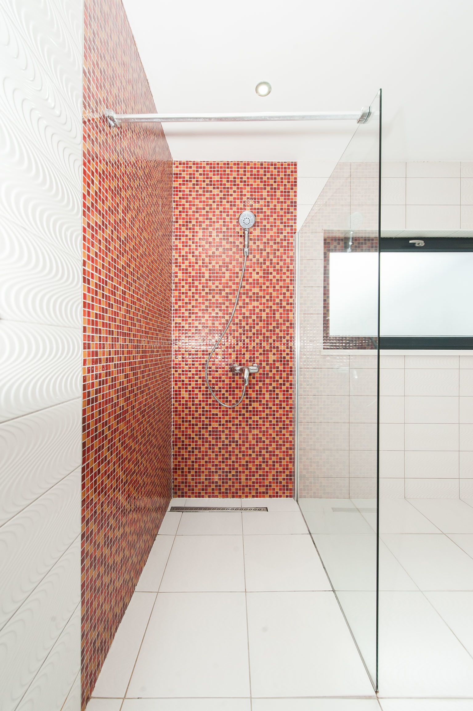 A modern bathroom with a shower area featuring red mosaic tiles, a glass shower door, and white tiled floors.