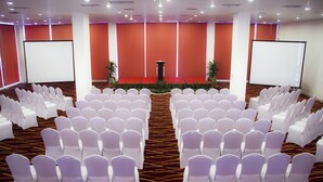 Un salón de conferencias con filas de sillas blancas, un atril y dos pantallas de proyección.
