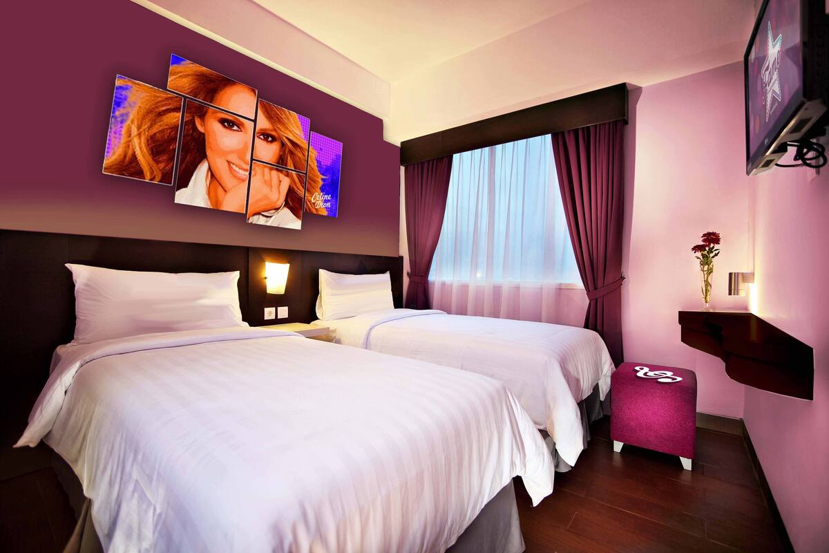 Quarto de hotel com duas camas, uma televisão grande fixada na parede, uma janela com cortinas e uma pequena mesa com um vaso.