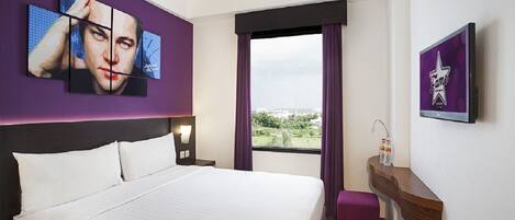 A hotel room with a large bed, a nightstand, a window with curtains, a wall-mounted TV, and a small wooden side table.