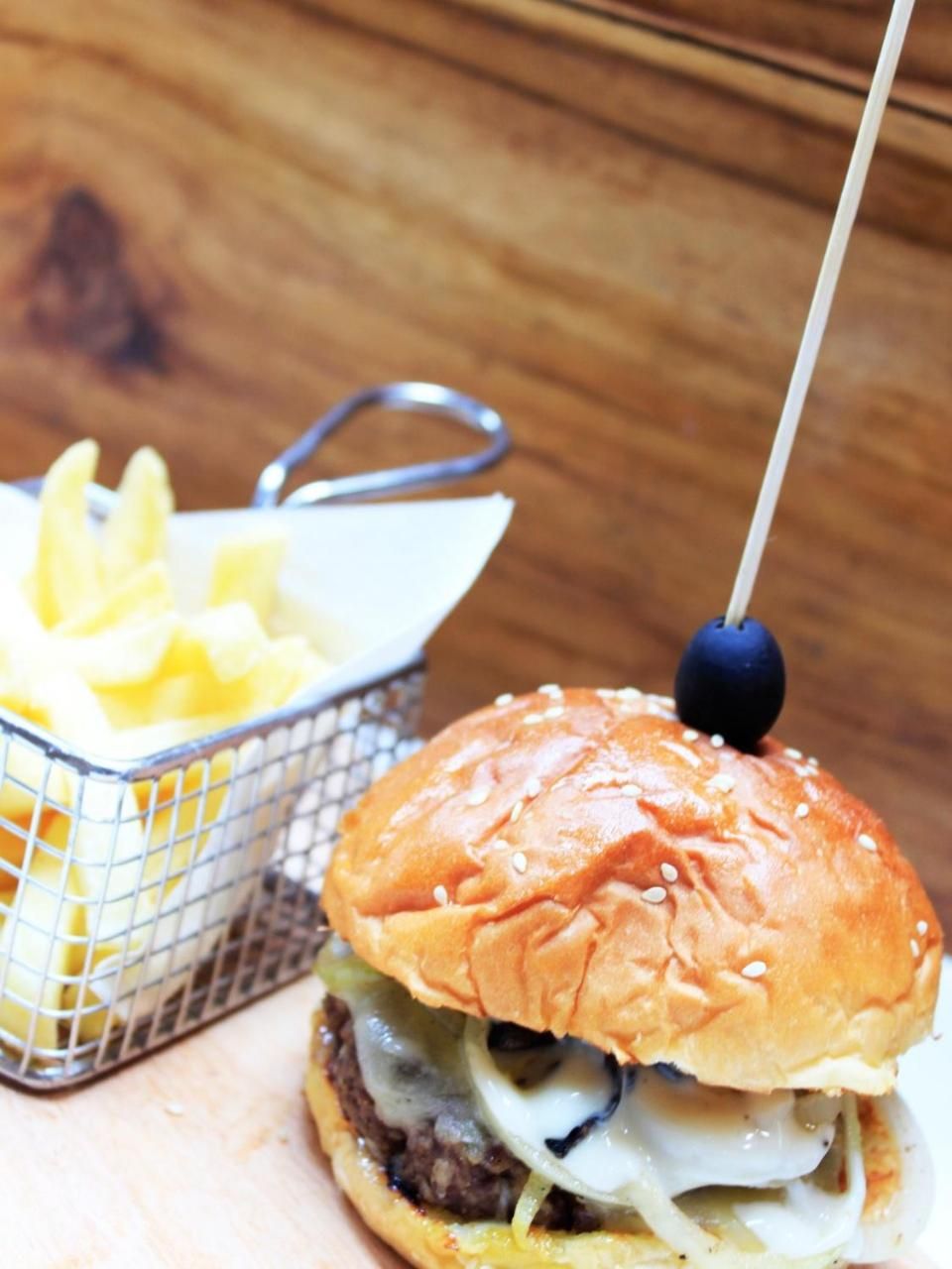 Een hamburger met sesamzaadbolletje en een blauwe prikker, vergezeld van een mandje friet.
