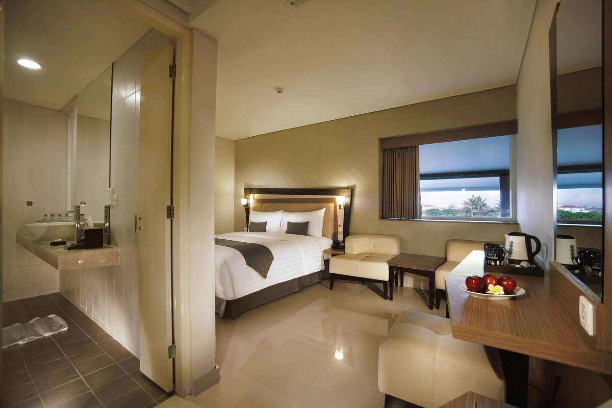 A hotel room with a bed, a desk with a laptop, a chair, a small table with a plate of fruit, a window with a view, and a bathroom with a sink and mirror.