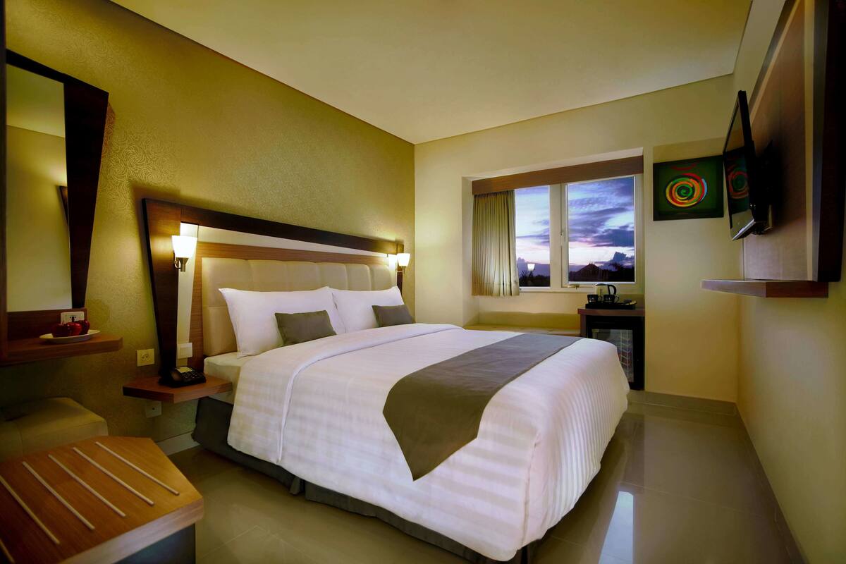 A hotel room with a large bed, bedside tables, a desk, a chair, a TV, and a window with a view of the sky.