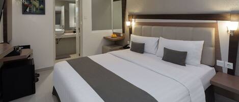 Une chambre d’hôtel avec un grand lit, une table de chevet avec une lampe, un miroir, une petite table avec un téléphone et des fruits, et un bureau avec un ordinateur et une imprimante.