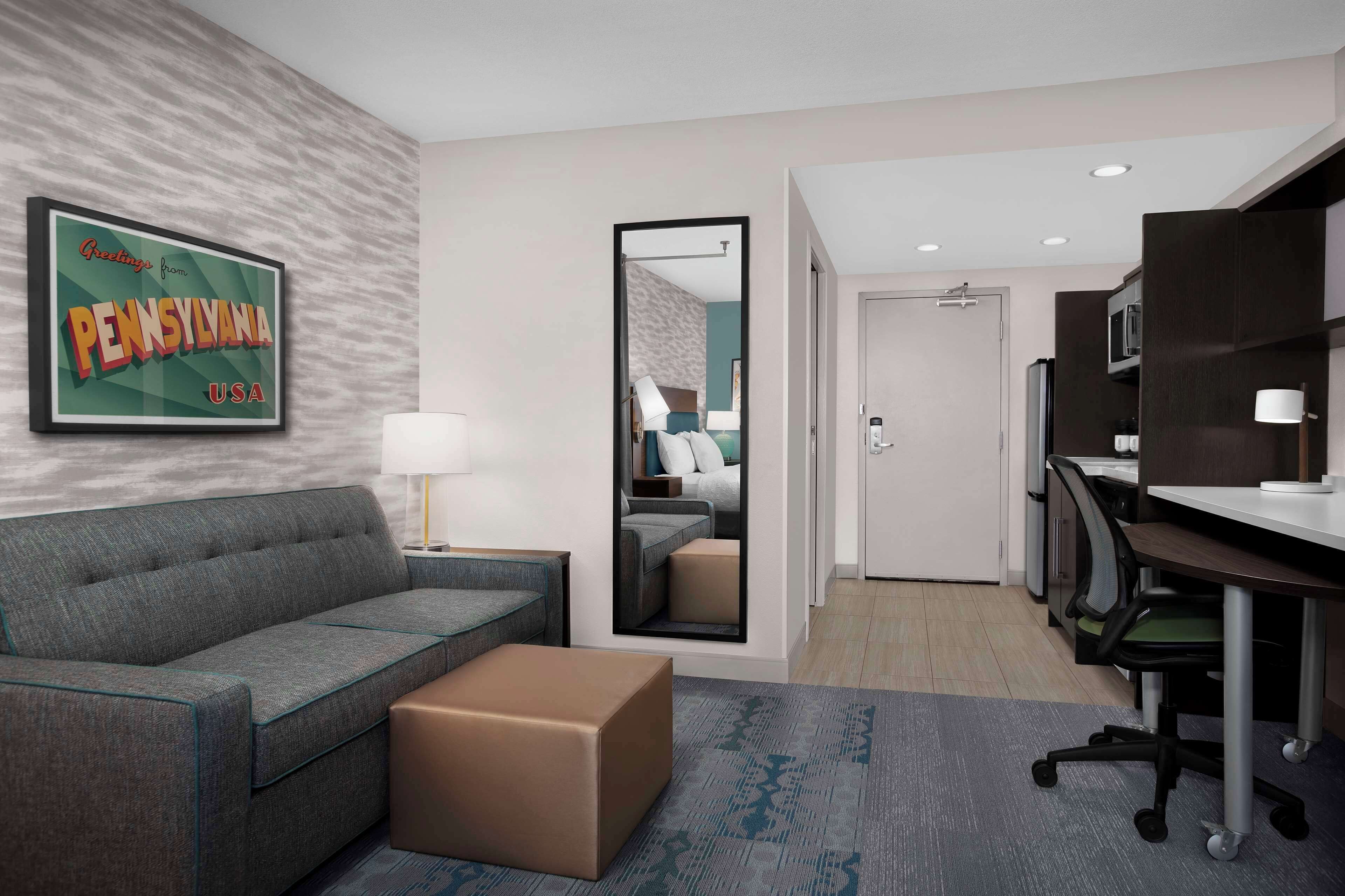 Una habitación de hotel moderna con un sofá gris, un pequeño otoman marrón, un escritorio con una silla, un espejo y un cartel enmarcado en la pared.