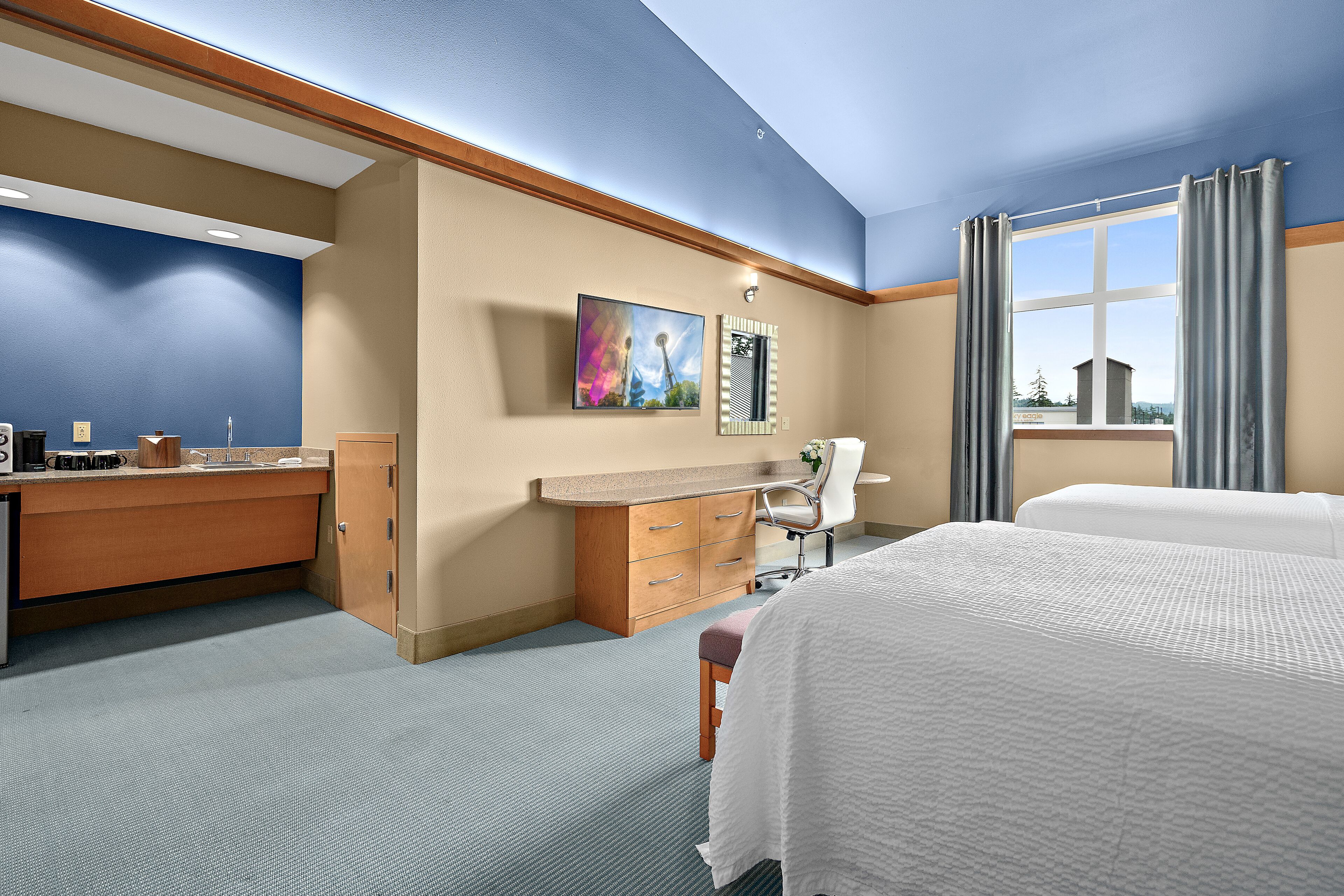 Habitación de hotel con dos camas, un escritorio con silla, una kitchenette pequeña y una ventana con cortinas.