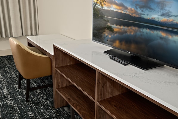 A modern hotel room with a large flat-screen TV displaying a scenic lake view, a wooden desk with a remote control, and a comfortable chair.