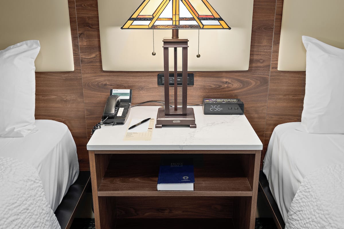 Habitación de hotel con dos camas, una mesita de noche con lámpara, reloj, teléfono, bolígrafo, Biblia y pared de paneles de madera.