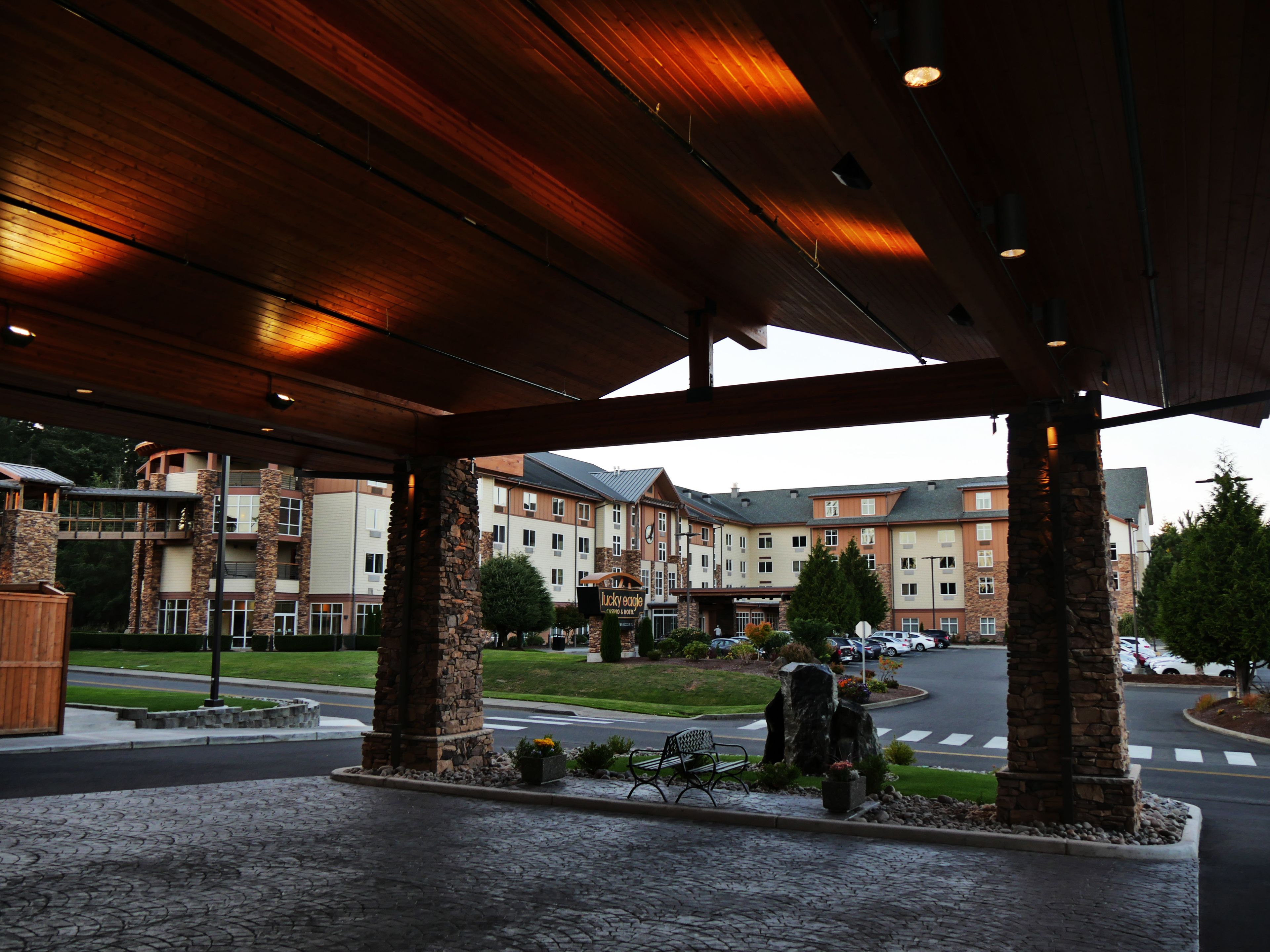 A covered outdoor area with stone pillars, a paved walkway, and a view of a hotel building in the background.