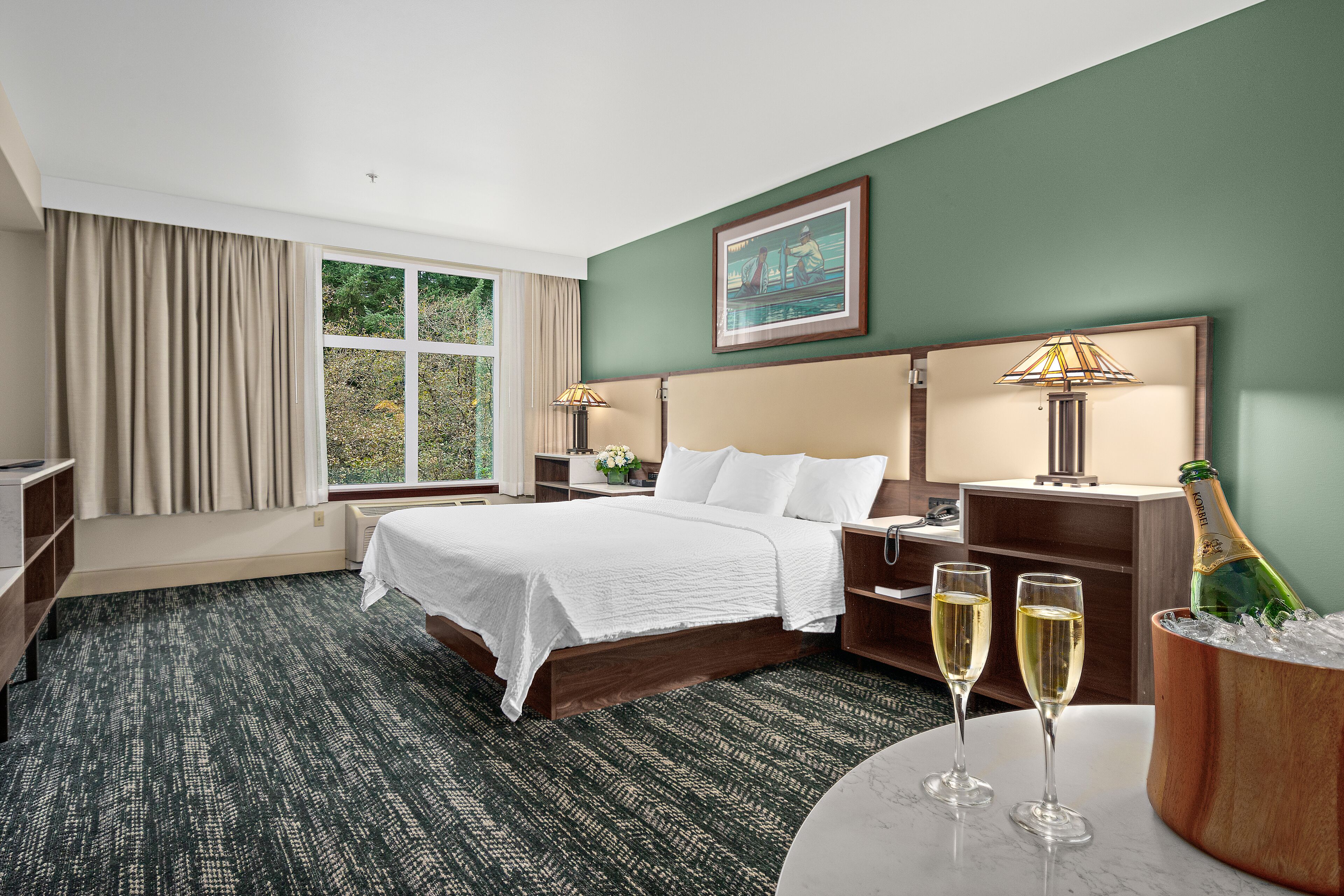 A hotel room with a bed, bedside tables, a nightstand, a lamp, a bottle of champagne, and two glasses of champagne on a table.