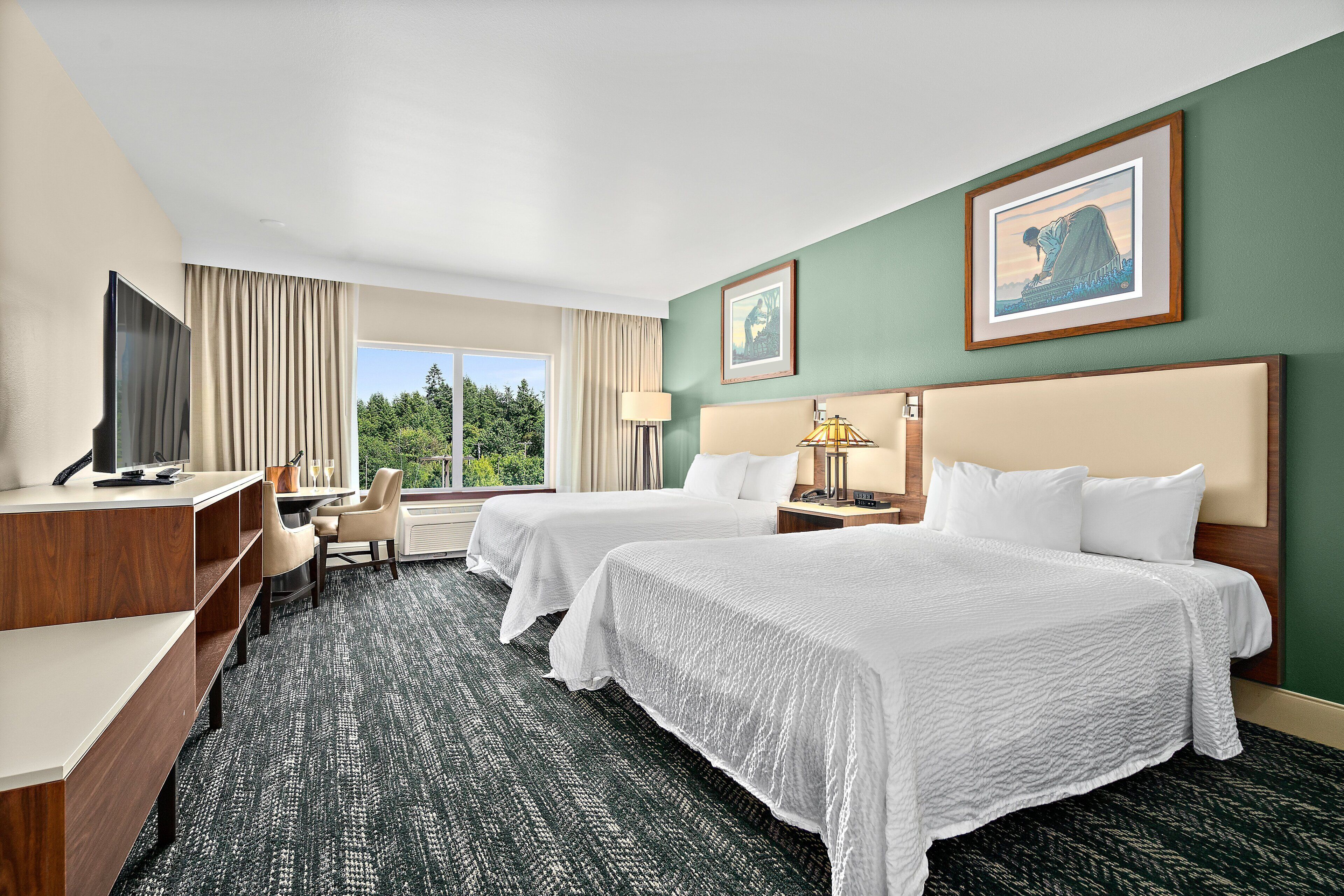 A hotel room with two beds, a desk, a chair, a TV, and a window with a view of trees.