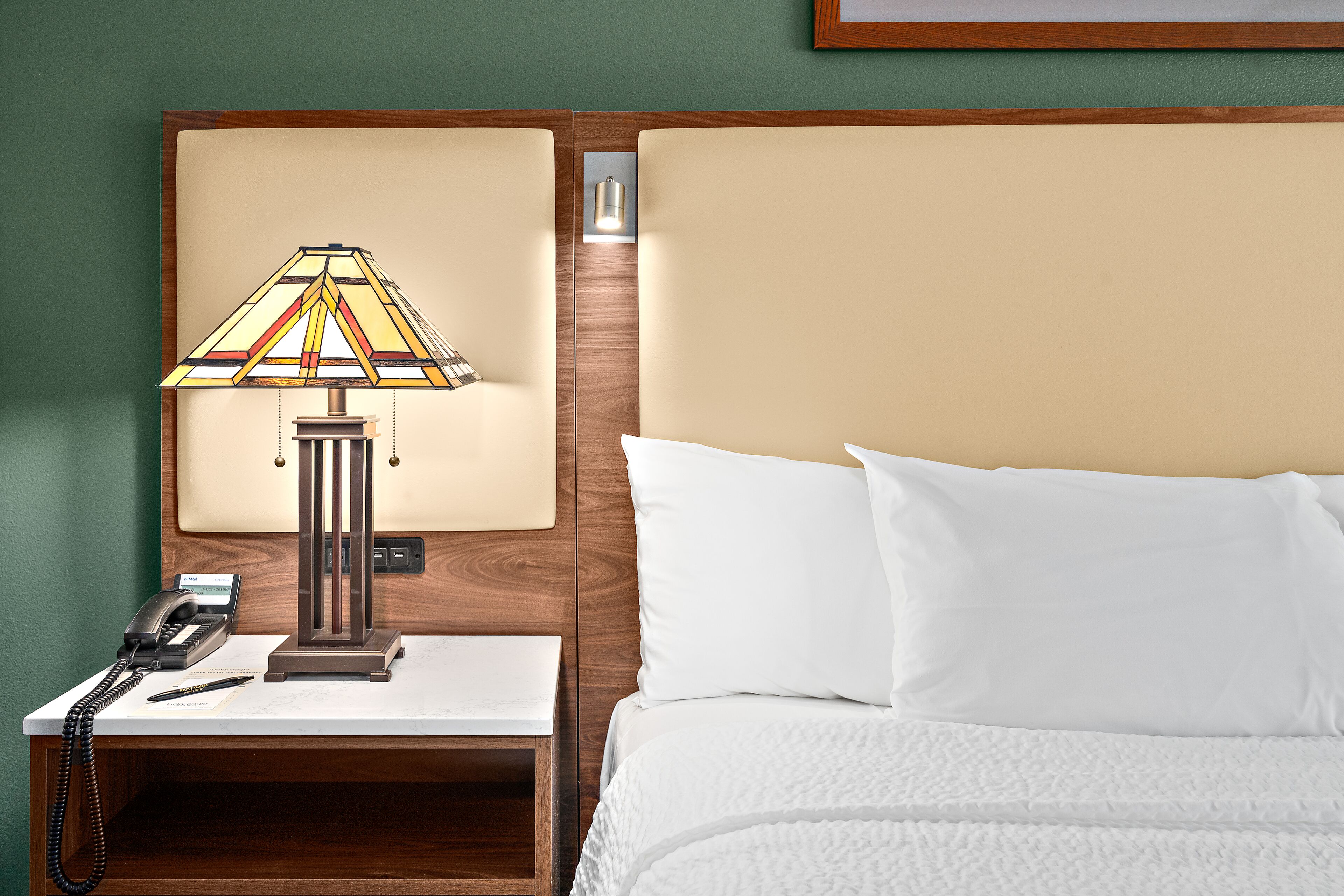 A hotel room with a bed, a nightstand with a lamp, a telephone, and a framed picture on the wall.